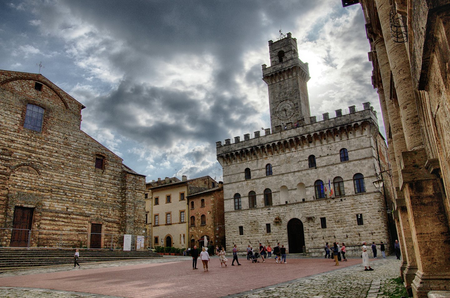 Persone passeggiano in una piazza storica con chiesa in mattoni e palazzo.